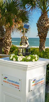 A polished trophy and floral centerpiece for “Constellation Furyk & Friends” stands on a beachside venue with palm trees and ocean behind it.