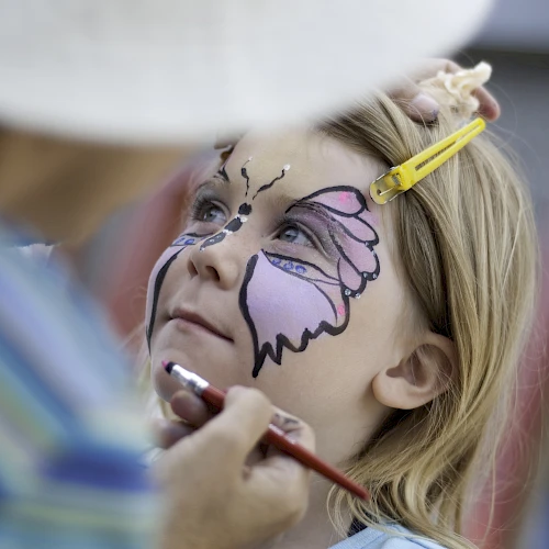 A child gets face-painted like a butterfly by an artist, with colors outlined in purple and pink, focused as makeup is applied.