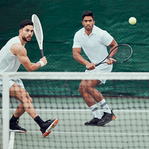 Two men in white shirts play doubles tennis at the net, ready to volley as a ball sails overhead.