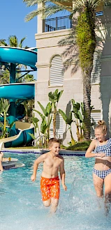 Kids and adults enjoy a sunny pool at a resort, with a blue water slide, palm trees, and a light-colored building backdrop.