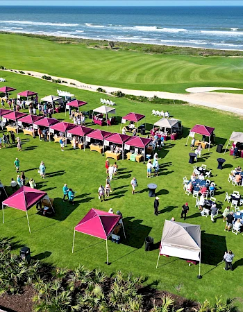 People gather under colorful tents on a large lawn by the ocean, enjoying an outdoor event with food and seating arrangements.
