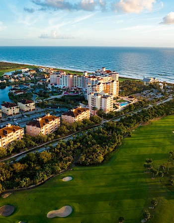 Aerial view of beachfront buildings, a golf course, and a body of water, with the ocean in the background under a blue sky.
