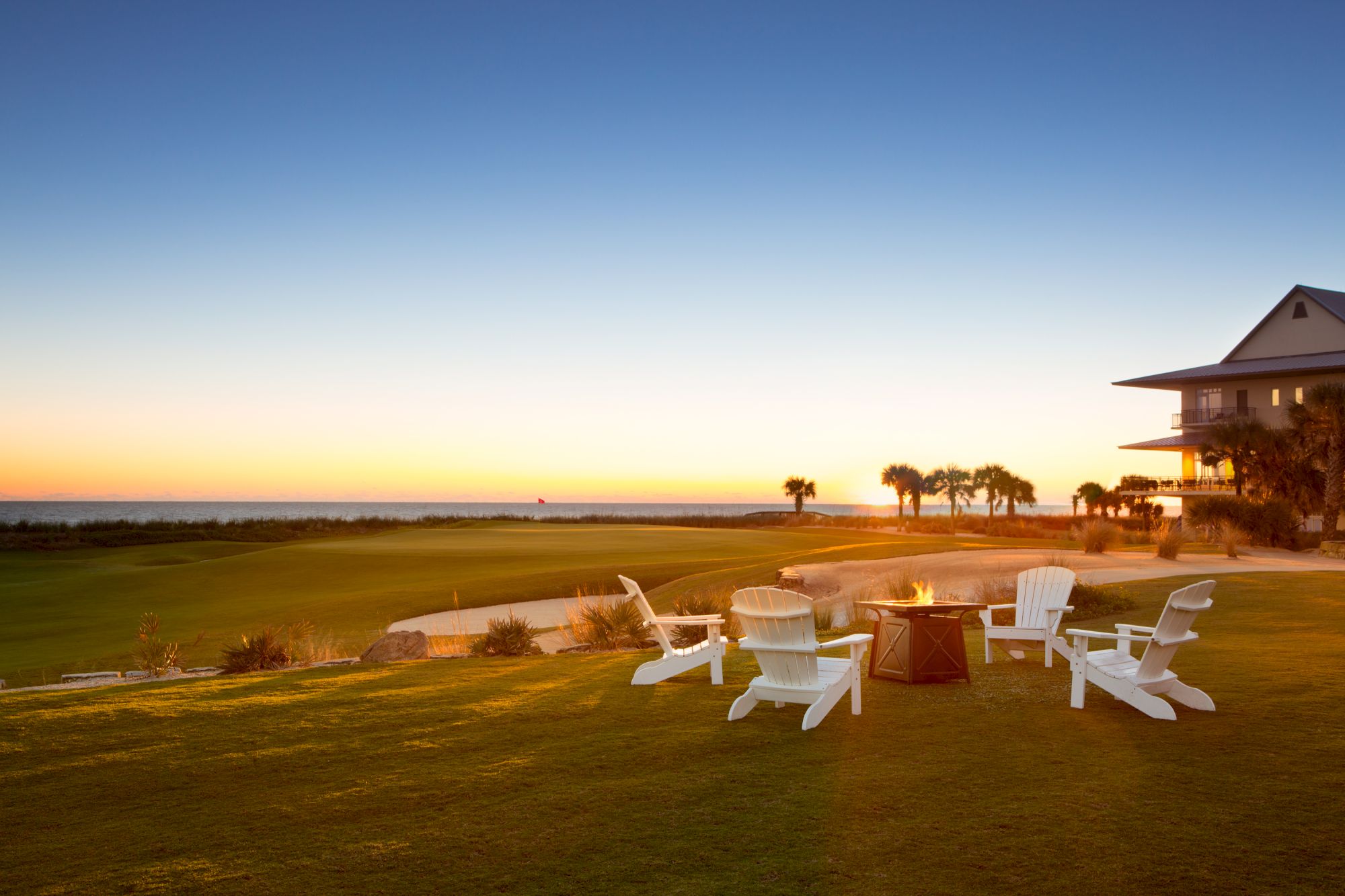 A scenic view of a golf course at sunset with a group of white chairs around a fire pit, near a building with palm trees in the background.