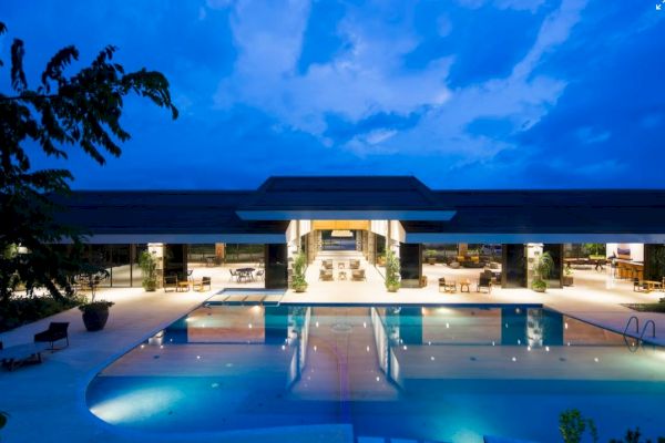 The image shows a luxurious, illuminated building with open spaces and a large reflecting pool, set against a twilight sky.