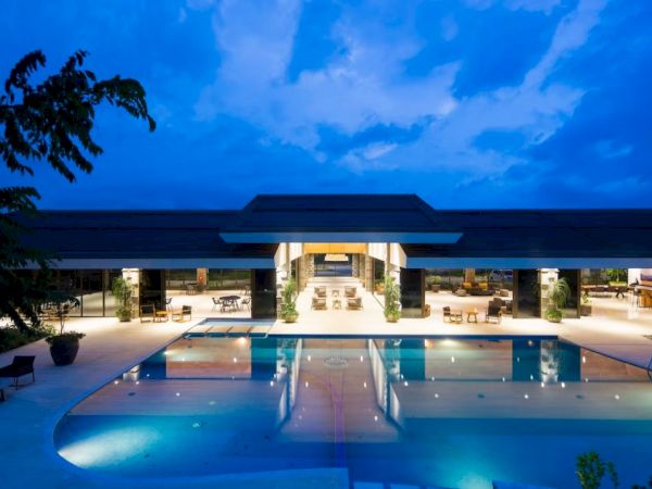 The image shows a luxurious, illuminated building with open spaces and a large reflecting pool, set against a twilight sky.