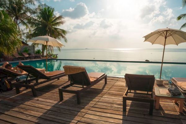 A serene poolside setting with lounge chairs, umbrellas, palm trees, and a view of the ocean under a partly cloudy sky.
