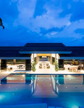 A luxurious building with an illuminated pool in the foreground and a modern structure in the background under a twilight sky.