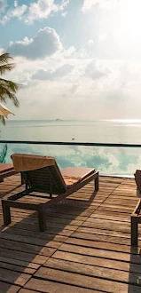 A serene poolside scene with lounge chairs, umbrellas, and palm trees, overlooking a calm sea under a partially cloudy sky.