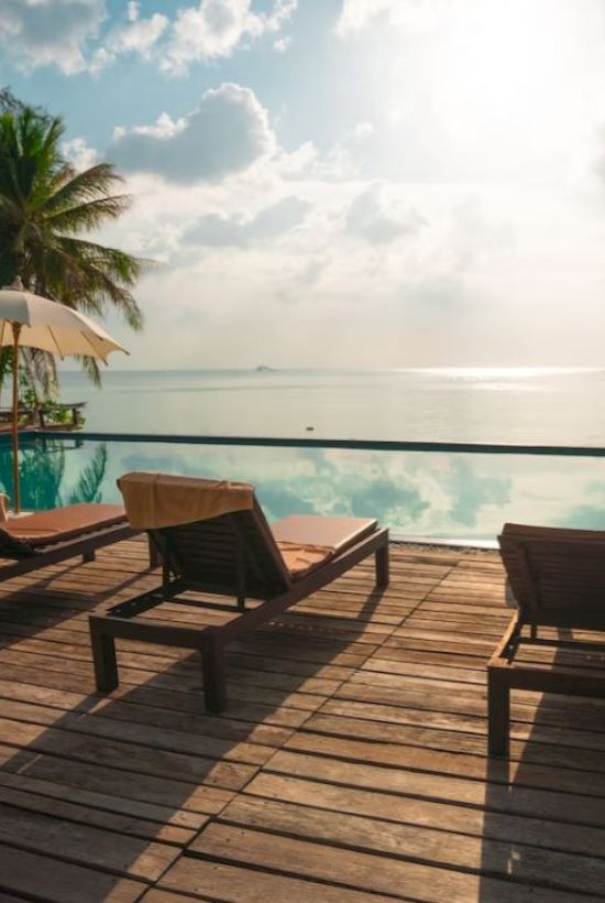 A serene poolside view with lounge chairs, umbrellas, and palm trees overlooking a calm ocean under a partly cloudy sky.