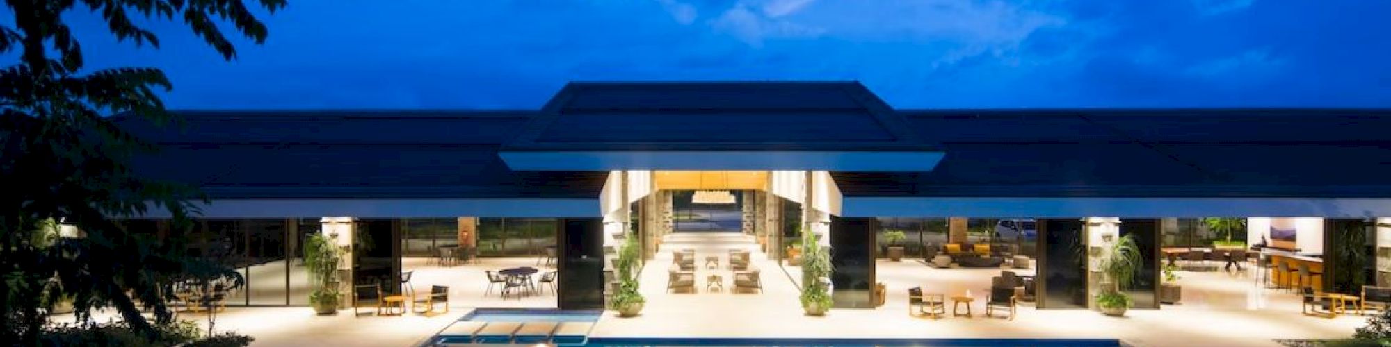 A luxurious pool area at dusk, featuring a modern open-air building with seating, illuminated under a vivid blue sky.