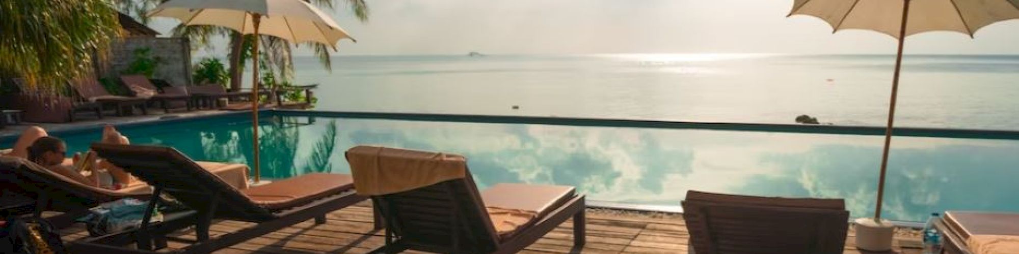 A serene poolside scene with lounge chairs, umbrellas, palm trees, and a clear view of the ocean under a bright sky.