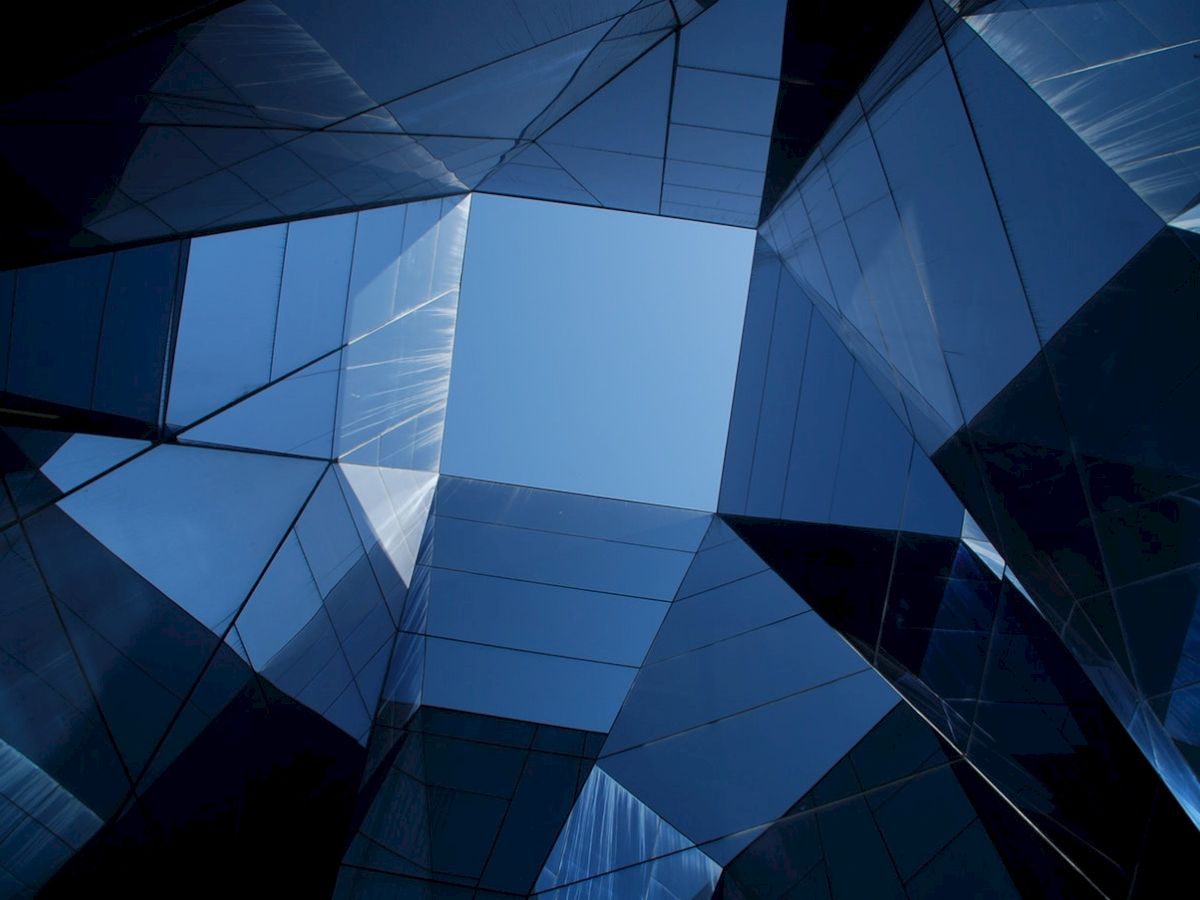 A geometric view upward through modern glass buildings with reflections and a blue sky in the center.