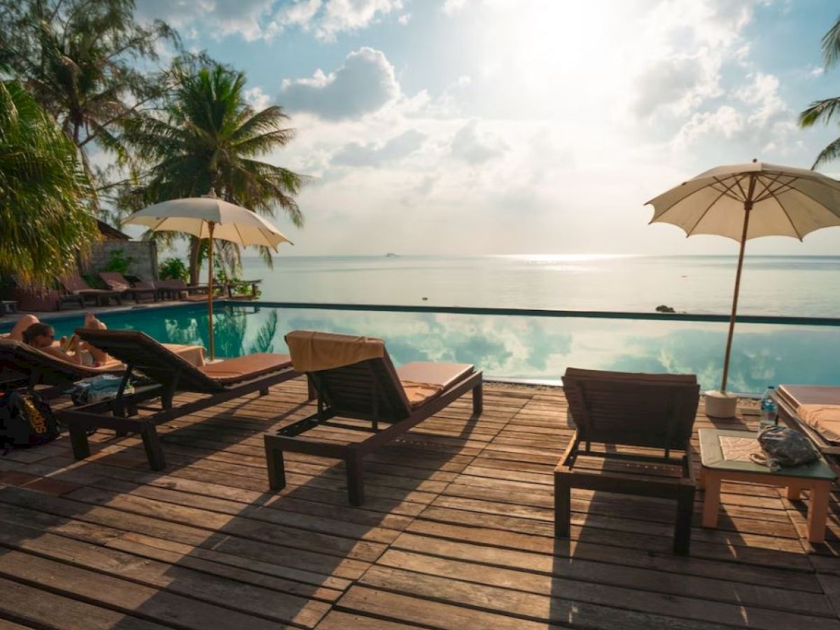 The image shows a tropical poolside with lounge chairs, umbrellas, and palm trees, overlooking a serene ocean view under a bright sky.