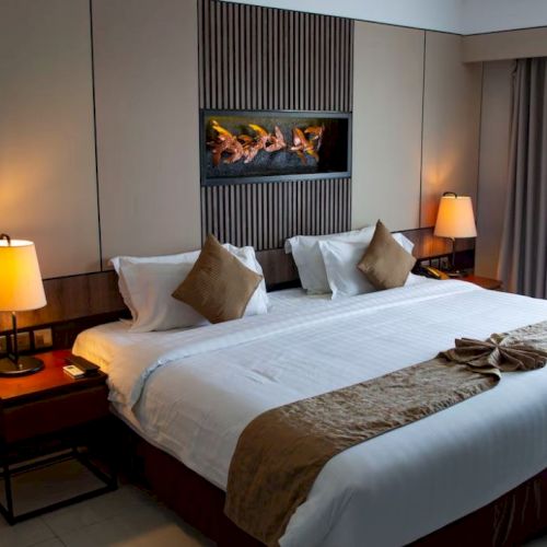 A hotel room with a large bed, decorative pillows, side lamps, a wall art piece, and a window with curtains softly lit from outside.