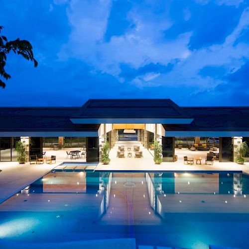 Luxurious building with open design, large illuminated pool, surrounded by trees under a twilight sky.