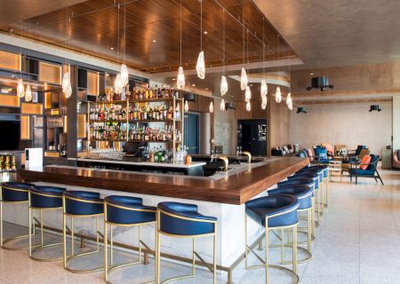 A modern bar with a sleek counter, blue chairs, hanging lights, and a wide assortment of bottles on shelves in an open, stylish space.