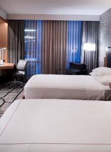 The image shows a modern hotel room with two beds, a desk, wall-mounted TV, and stylish decor, featuring patterned wallpaper and curtains.