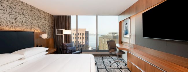 A modern hotel room with a large bed, wall-mounted TV, desk and chair, and a window view of buildings and water.