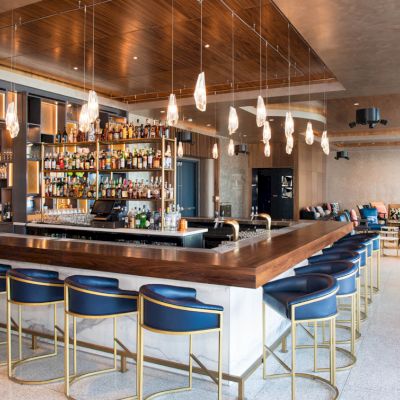A modern bar with a wooden counter, blue chairs, pendant lights, and a stocked back shelf; seating area in the background.