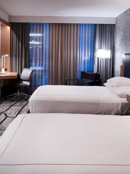 The image shows a modern hotel room with two double beds, a wall-mounted TV, a desk, and a window with curtains, and stylish decor.