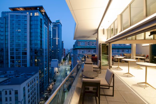 A modern balcony overlooks a city street at dusk, with tables and chairs set up along the outdoor space.
