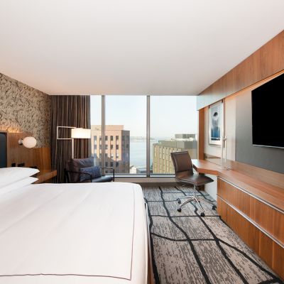 A modern hotel room with a bed, TV, desk, and large window offering a cityscape view, featuring stylish decor and furnishings.