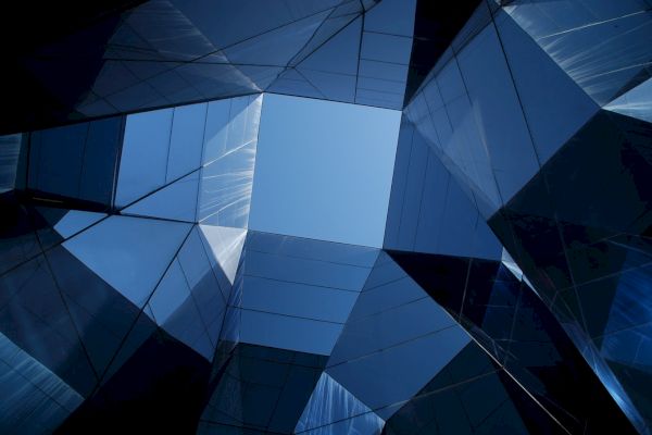 The image shows a view up through modern geometric glass architecture, with a clear blue sky visible through the center.