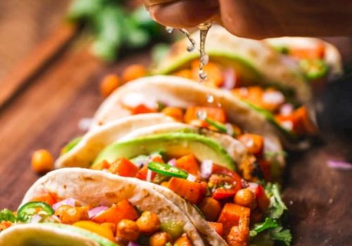 A hand squeezes lime over a row of tacos with chickpeas, avocado, and vegetables, garnished with cilantro, on a wooden board.