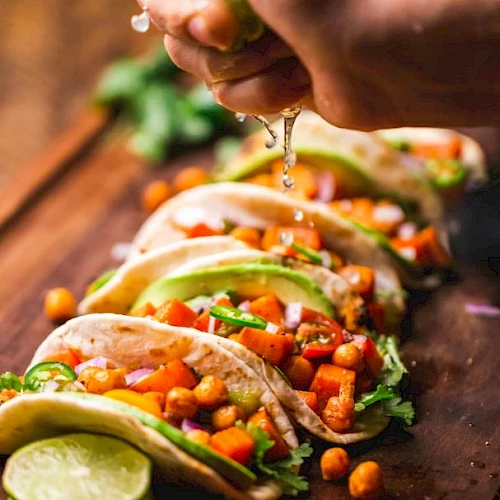A hand squeezes lime over a row of tacos with chickpeas, avocado, and vegetables, garnished with cilantro, on a wooden board.