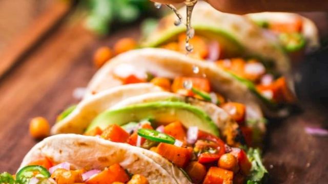 A hand squeezes lime over a row of tacos with chickpeas, avocado, and vegetables, garnished with cilantro, on a wooden board.
