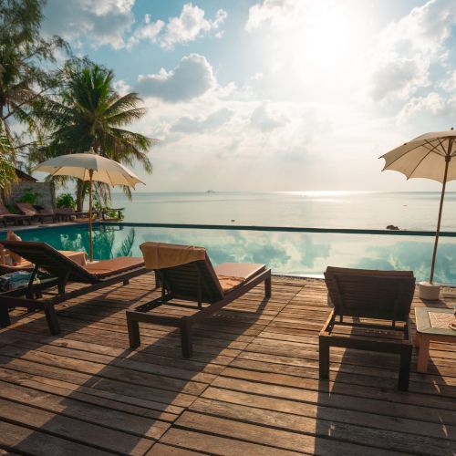 A serene beach scene with lounge chairs, umbrellas, palm trees, and an infinity pool overlooking a calm ocean under a sunny sky.