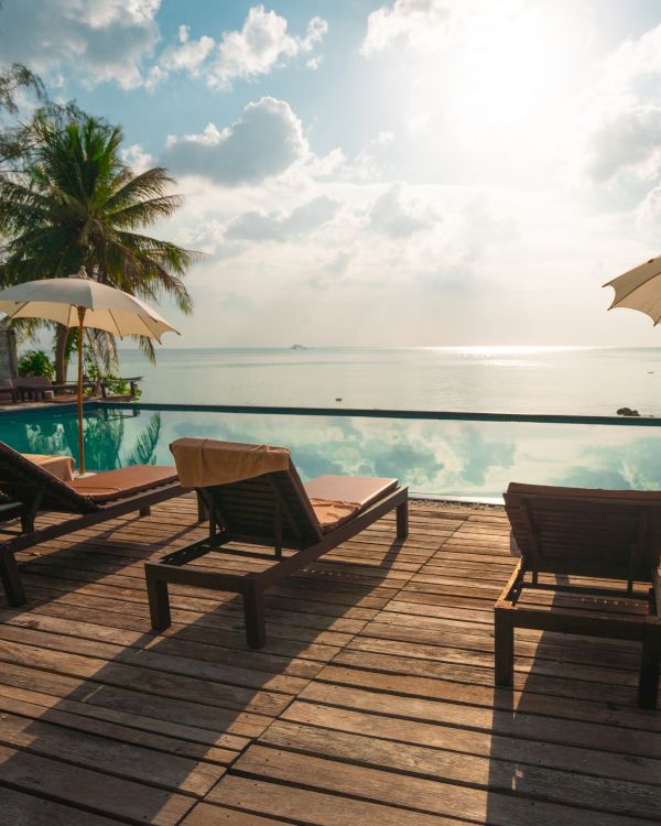 A serene beach scene with lounge chairs, umbrellas, palm trees, and an infinity pool overlooking a calm ocean under a sunny sky.