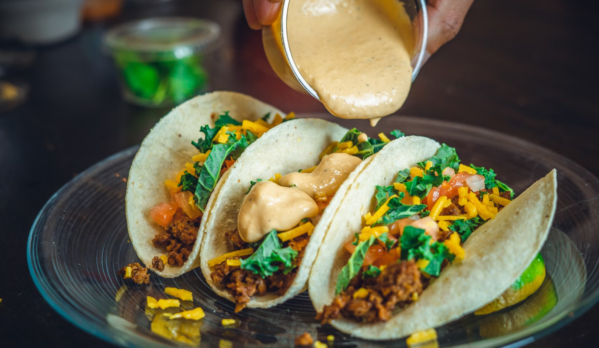 Three tacos with toppings like lettuce, cheese, and sauce being poured, arranged on a plate.