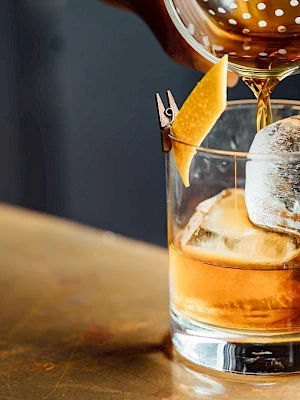 A drink is being poured over large ice cubes in a glass with a lemon garnish, set on a wooden surface.