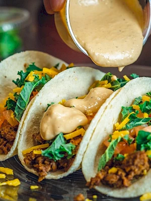 Three tacos filled with ground meat, shredded cheese, and greens on a plate, with sauce being poured over them.