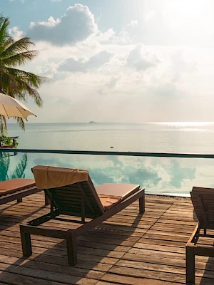 Poolside scene with lounge chairs, umbrellas, and an ocean view, framed by palm trees under a bright sky with clouds.