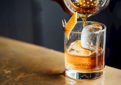 A hand pours a brown cocktail into a glass with ice and a citrus twist on a metal surface.