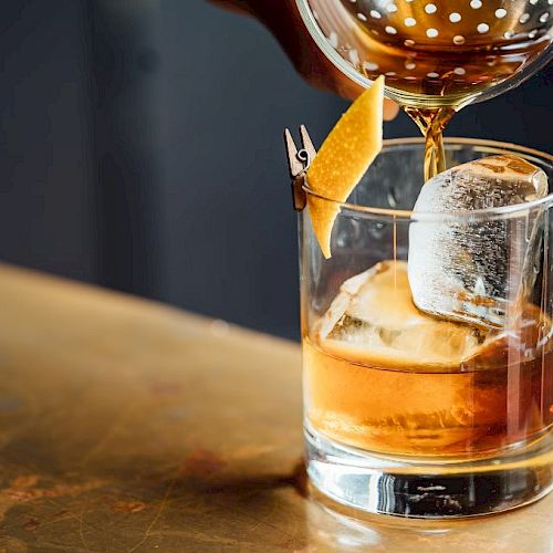 A hand pours a brown cocktail into a glass with ice and a citrus twist on a metal surface.