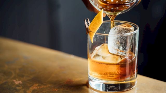 A hand pours a brown cocktail into a glass with ice and a citrus twist on a metal surface.