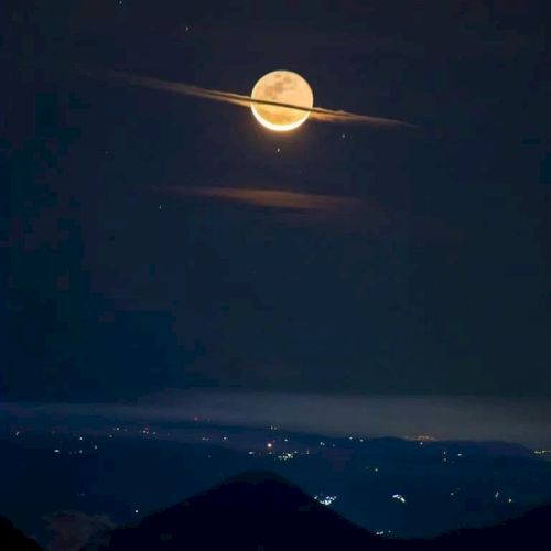 A full moon with clouds resembling Saturn's rings appears over a landscape with distant city lights against a dark sky.