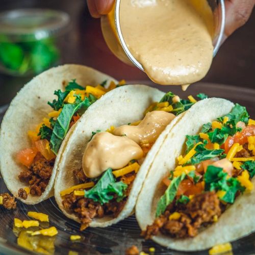 Three tacos filled with meat, cheese, lettuce, and tomatoes are on a plate, with sauce being poured on top at the end.