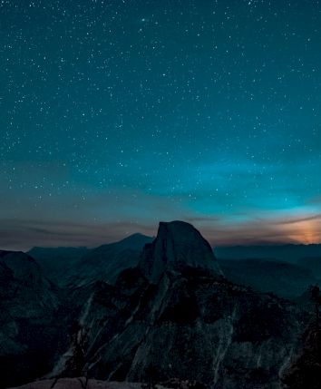 A starry night sky over a mountain landscape with a glowing horizon in the background.