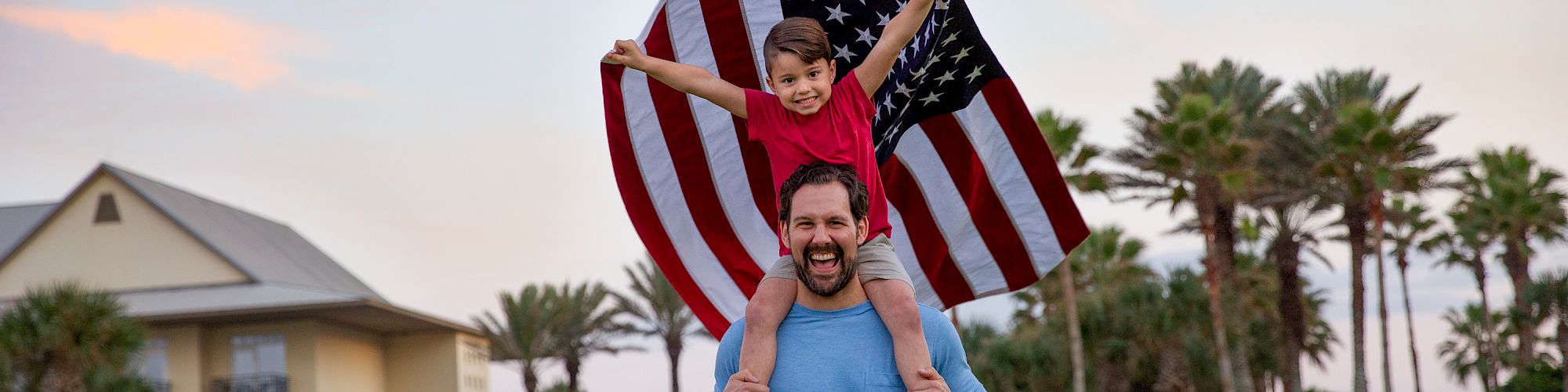 A man carries a child on his shoulders, with the child holding an American flag, surrounded by palm trees and a house in the background.