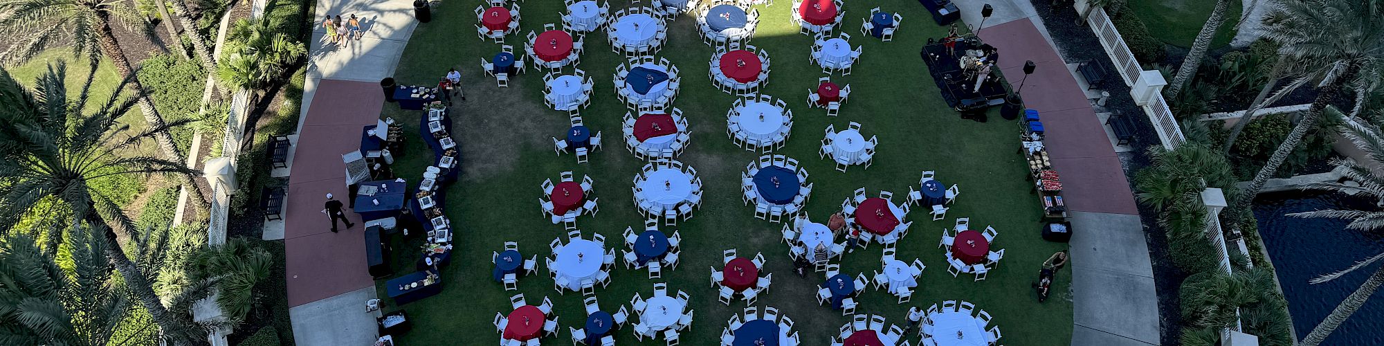 Aerial view of an outdoor event with round tables and chairs arranged on a grassy area, surrounded by palm trees and pathways.