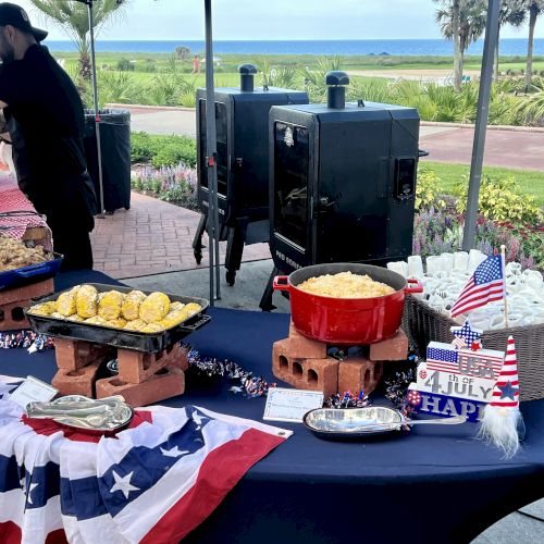 A festive outdoor setup with food, American flags, a "4th July" sign, grills, and a person preparing food, likely for a celebration.