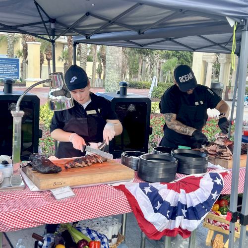 Two people preparing and serving barbecue food under a tent, with a table decorated in a patriotic theme.