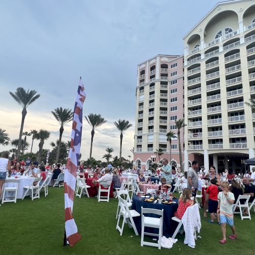 Outdoor gathering on a lawn with people sitting at tables and a tall building in the background, surrounded by palm trees.