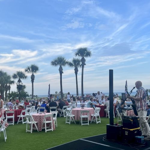 Outdoor event with people seated at tables under palm trees, near a speaker on a microphone.