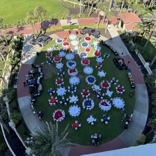 An outdoor event with round tables covered in red, white, and blue tablecloths, surrounded by palm trees and greenery.