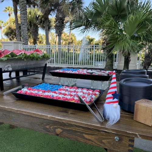 A picnic table with an American flag-themed dessert, fruits, and gnome decorations in an outdoor setting with palm trees.